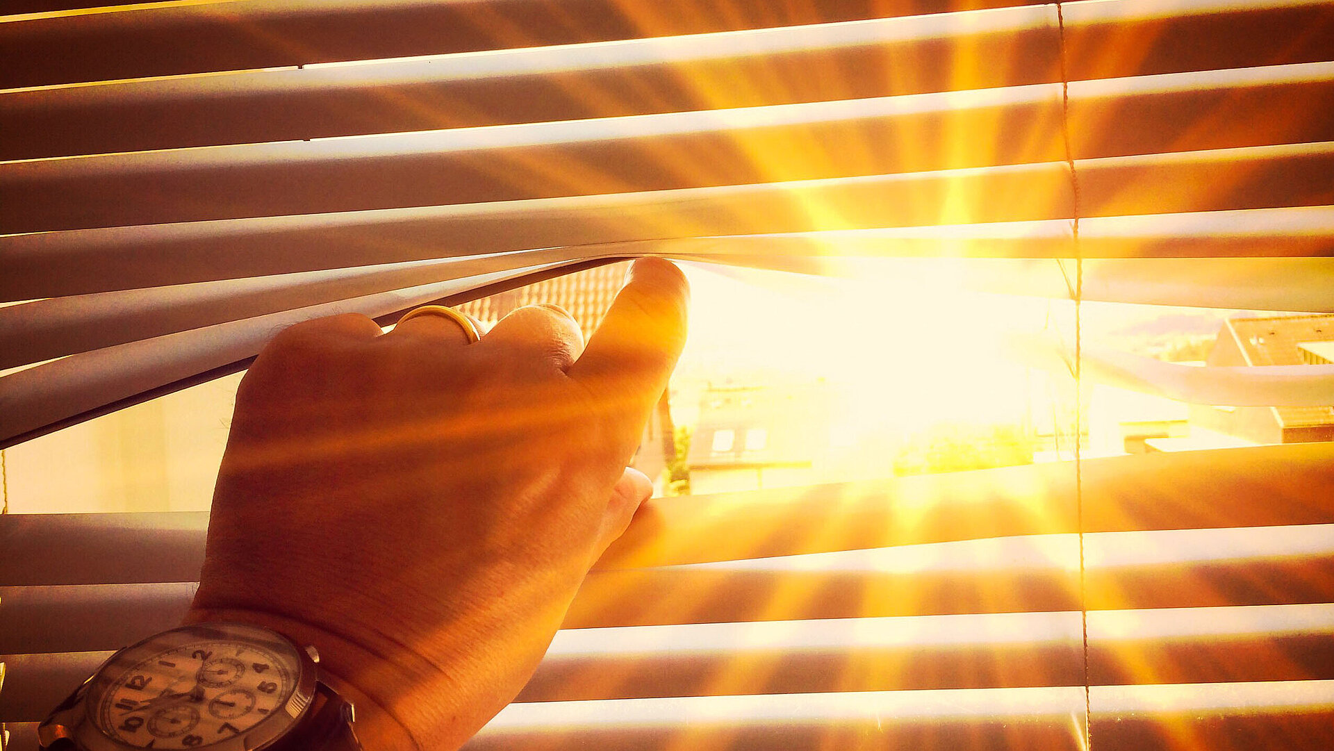 Eine Hand öffnet einen Spalt in einer Jalousie, durch das dahinterliegende Fenster scheint die Sonne