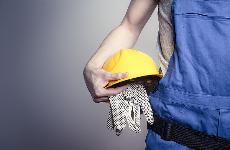 Eine Person in blauer Latzhose hält einen gelben Sicherheitshelm und Schutzhandschuhe unter den Arm geklemmt. Das Bild steht symbolisch für den Bereich Arbeitsschutz und Schutzausrüstung.