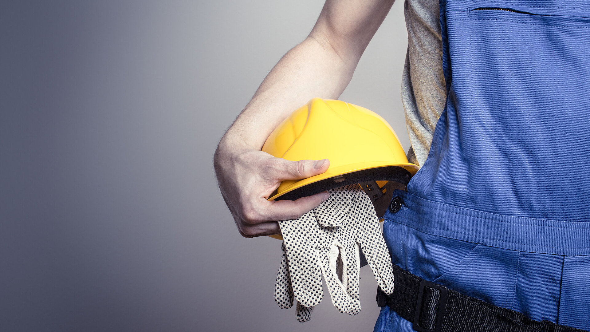 Eine Person in blauer Latzhose hält einen gelben Sicherheitshelm und Schutzhandschuhe unter den Arm geklemmt. Das Bild steht symbolisch für den Bereich Arbeitsschutz und Schutzausrüstung.