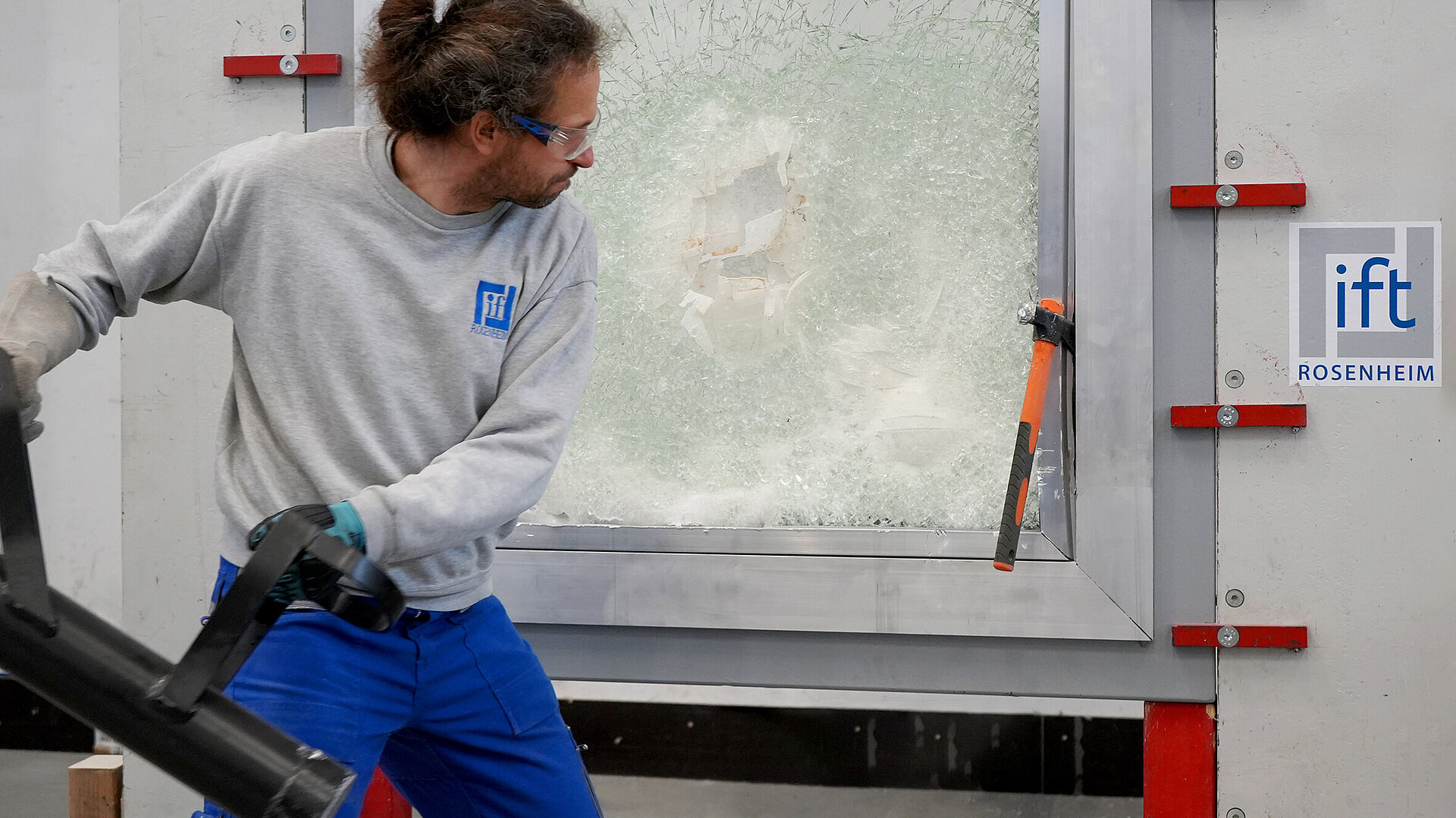 ift-Prüfer mit Ramme in der Hand steht vor einem Fenster mit gebrochener Scheibe zur Vandalismusprüfung