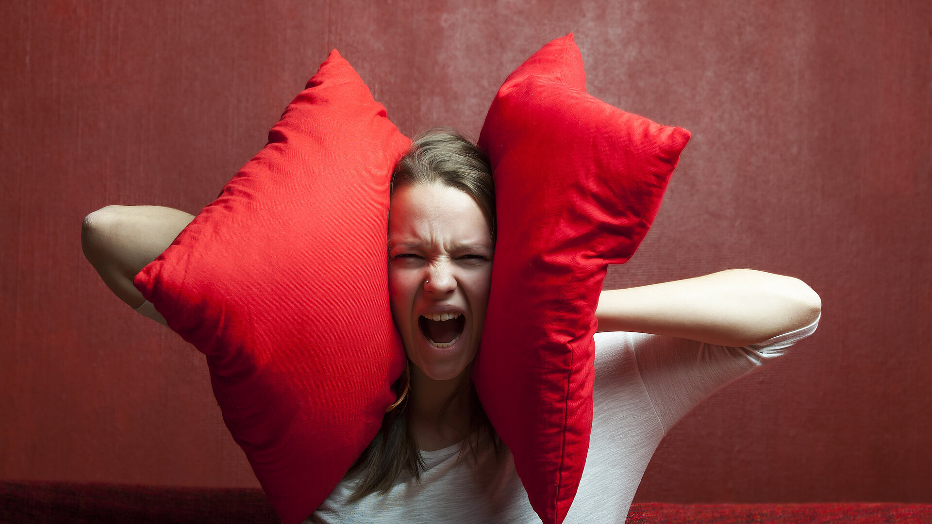 Eine schreiende Frau hält sich zwei rote Kissen an die Ohren. Das Bild steht symbolisch für Schallschutz