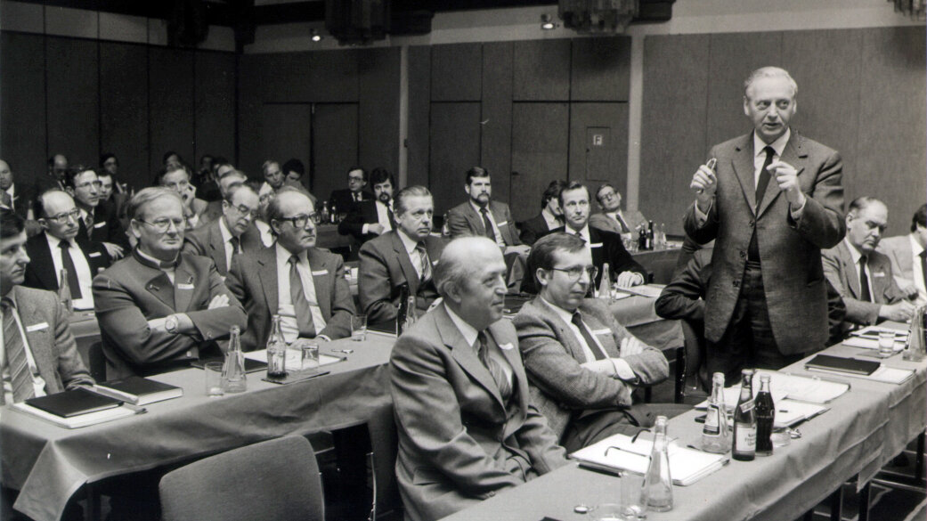 Schwarz-Weiß-Bild von einer Gruppe an Menschen, die in einem Vortragssaal an Tischen sitzen, eine Person steht und spricht